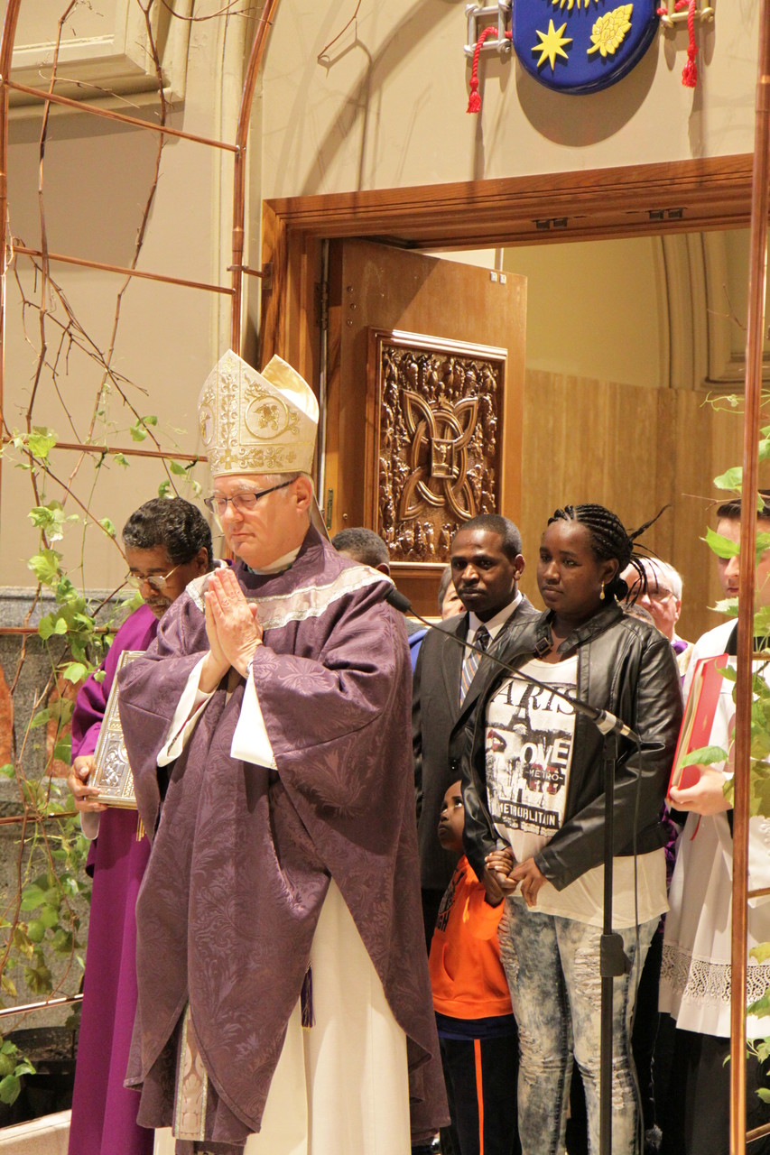 In a gesture symbolic of the opening of God’s love and forgiveness during the Year of Mercy, Bishop Thomas J. Tobin opened the Holy Door at the Cathedral of SS. Peter and Paul during a ceremony on Sunday. As the bishop stops to pray after passing through the doorway, he is joined by Ibrahim Aray, Sambtu Mencha and their four children. The Eritrean family, now living in Providence, came to Rhode Island several years ago as refugees.