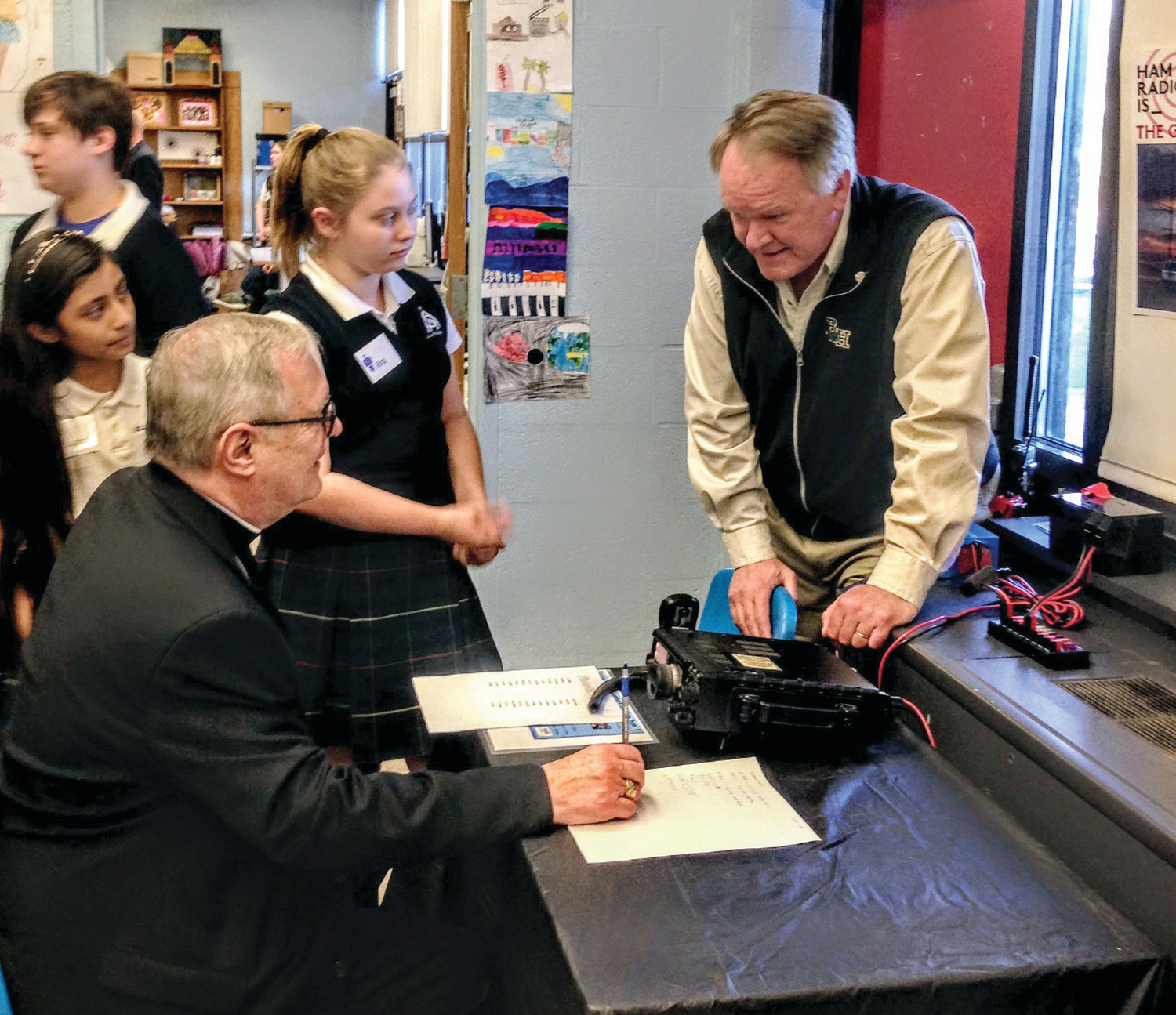 Bishop Thomas J. Tobin attends the annual STEAM Night and gets a pointer from Newport amateur radio/CyberPatriot coach Mike Cullen before getting on the air and seeing “communication resiliency” at work.  Students Arianna, left, and Ava stand ready to assist in making the contact.