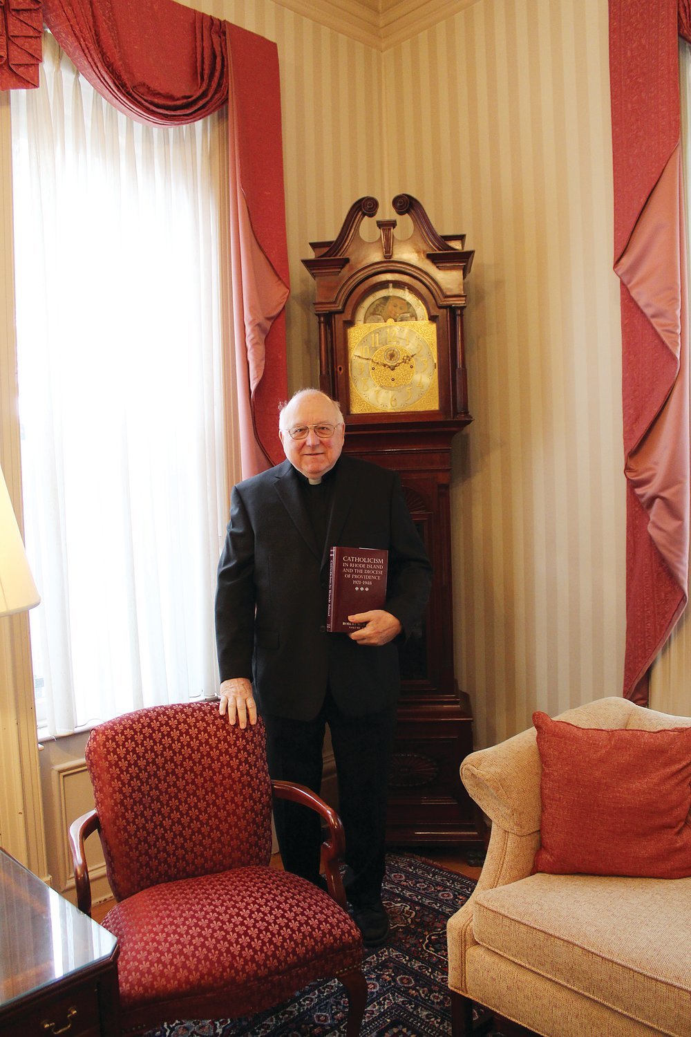 DOCUMENTING THE PASSAGE OF TIME: Father Robert W. Hayman holds a copy of his latest work in his “Catholicism in Rhode Island” series. The 600-page volume, exhaustive in its research, offers a panoramic history of the diocese from 1921-1948.
